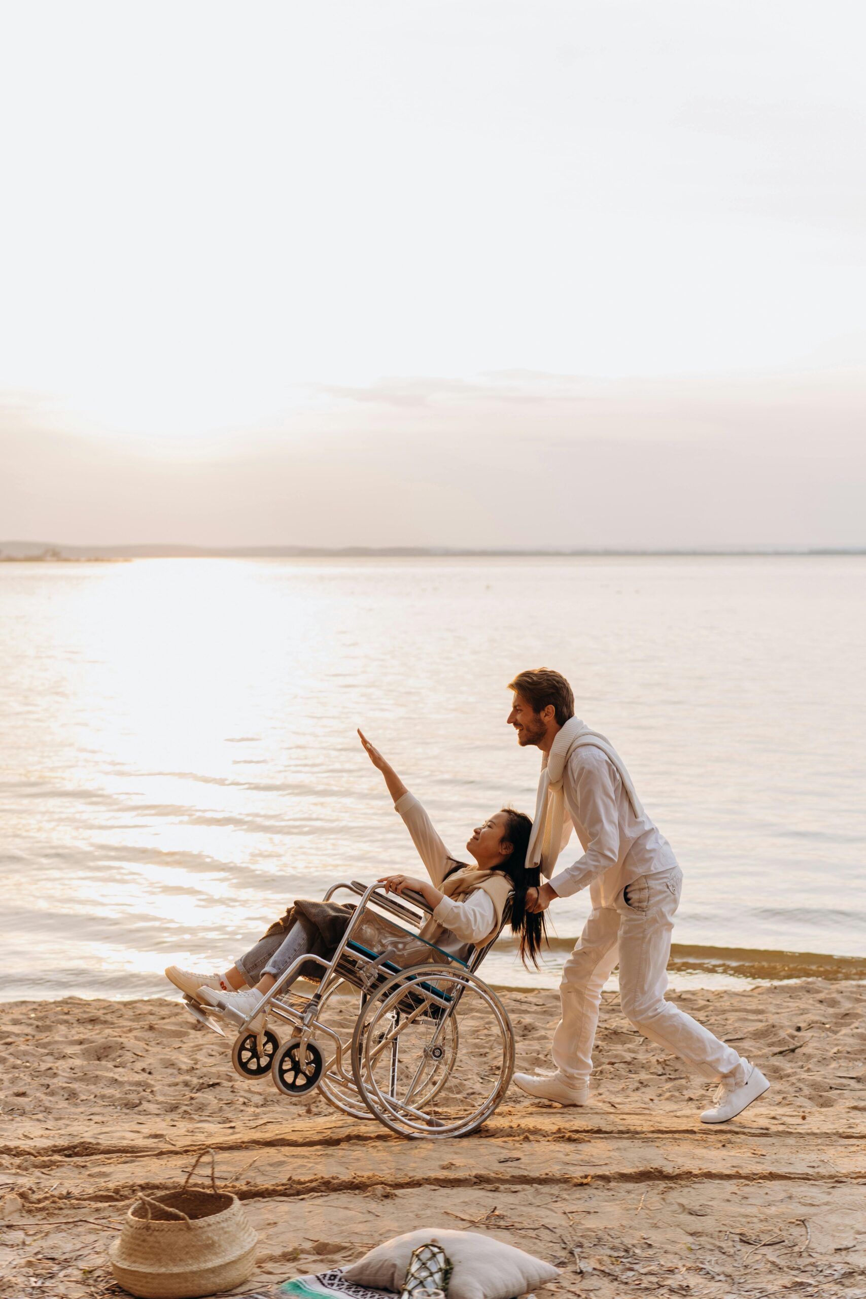 A couple enjoys a romantic moment on the beach, with a man pushing a woman in a wheelchair towards the sunset.