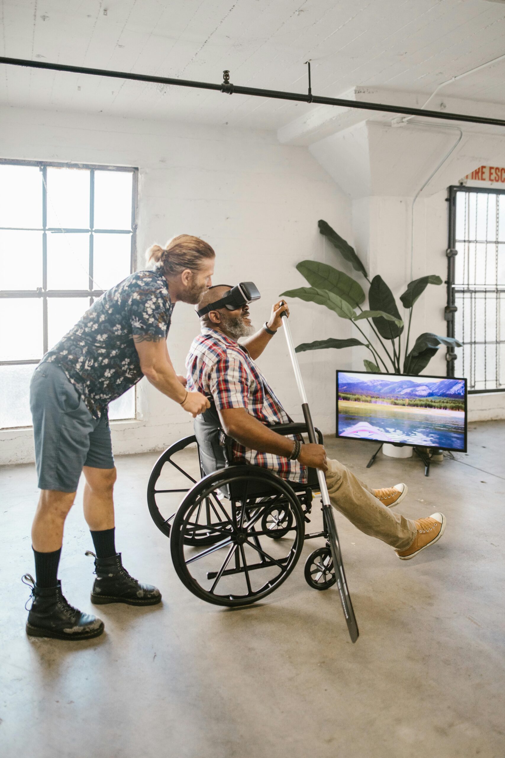 Man assisting another in a wheelchair using a VR headset indoors.