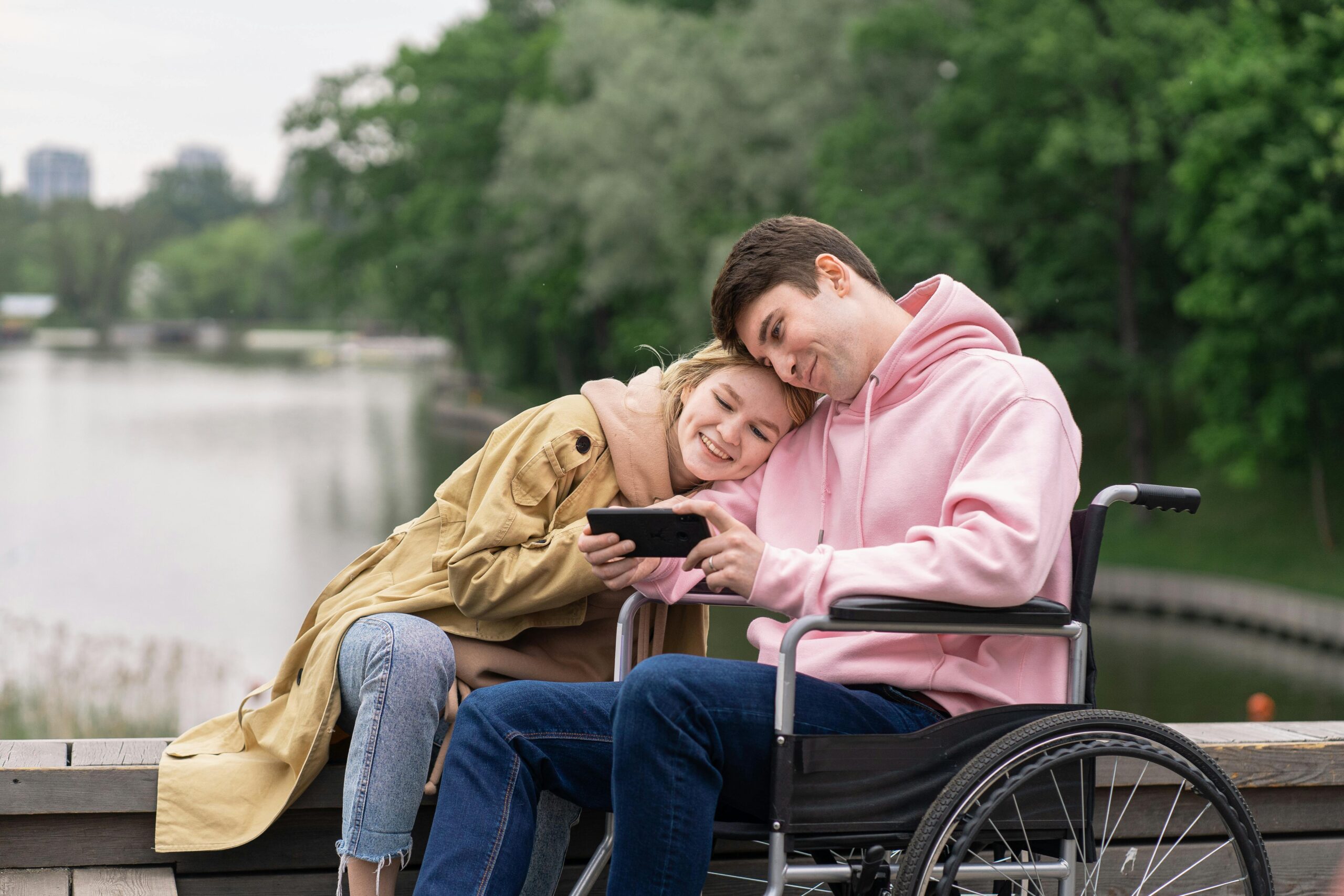 Smiling couple by lakeside enjoying a moment on smartphone, one in a wheelchair.